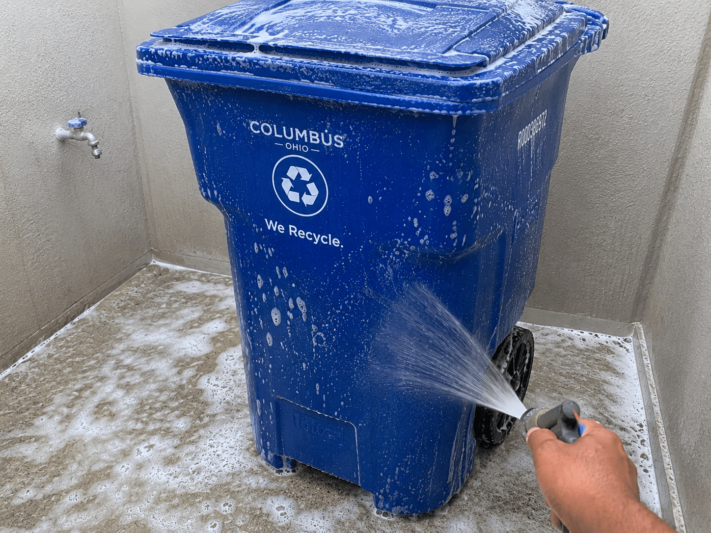 Hand spraying water to rinse soap suds off a blue Columbus Ohio recycling bin.