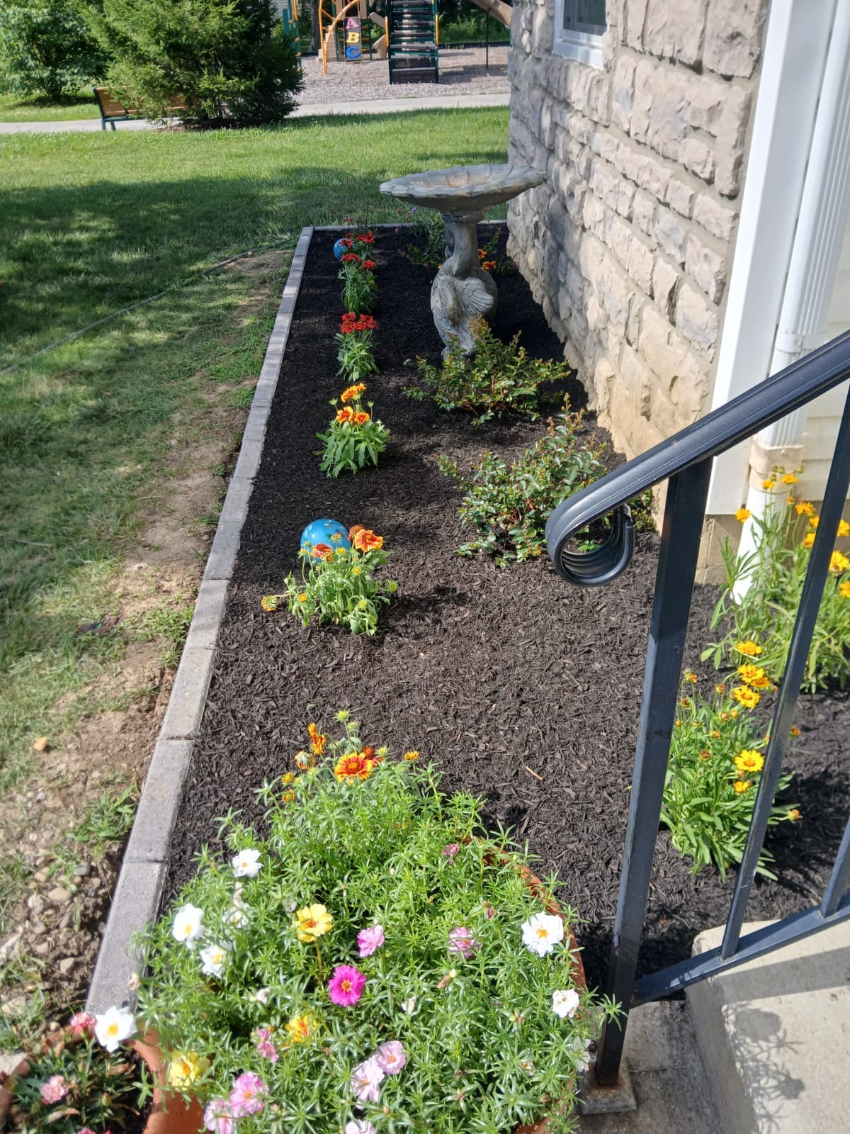 Colorful flowers and a swan birdbath in a dark mulched garden bed beside a stone wall.