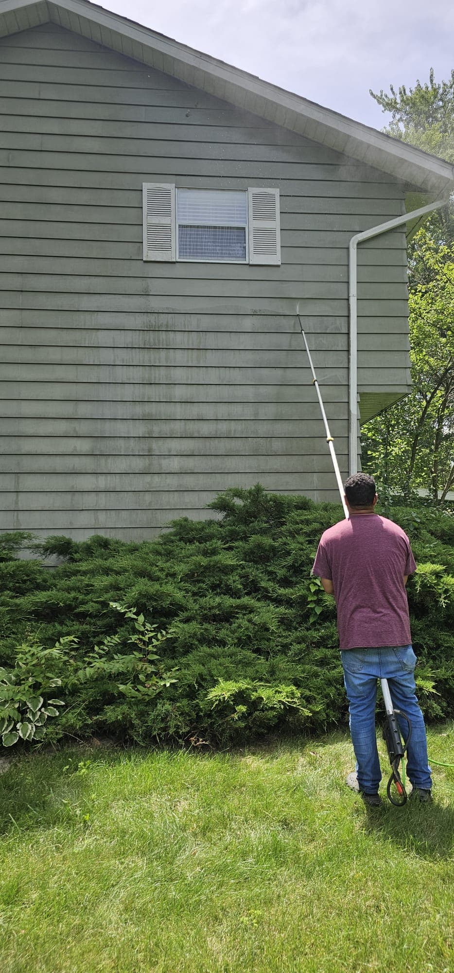 Man uses a long extension wand to pressure wash dirty green siding on a house.