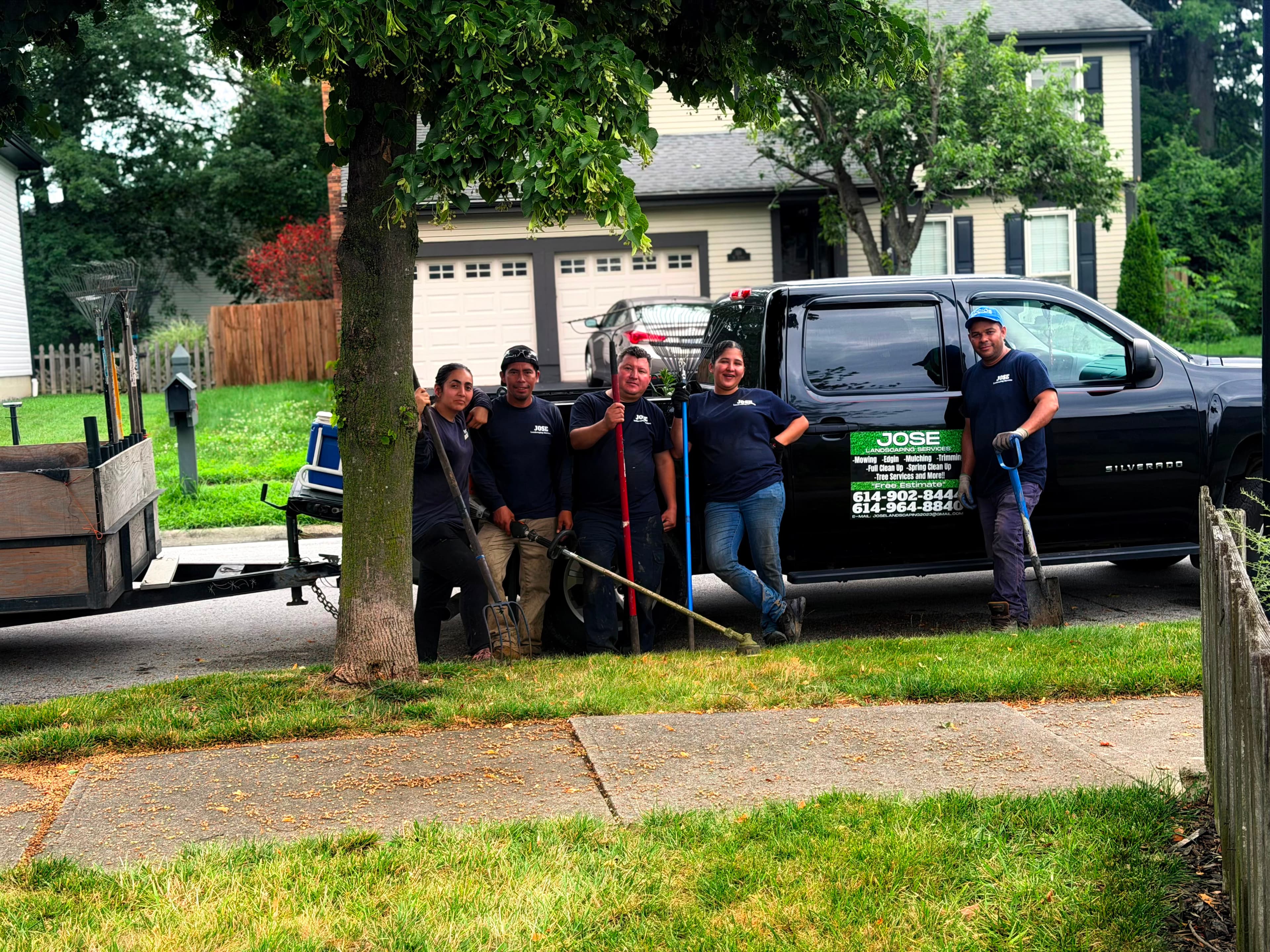 Five landscapers pose with tools in front of a black pickup truck and equipment trailer.