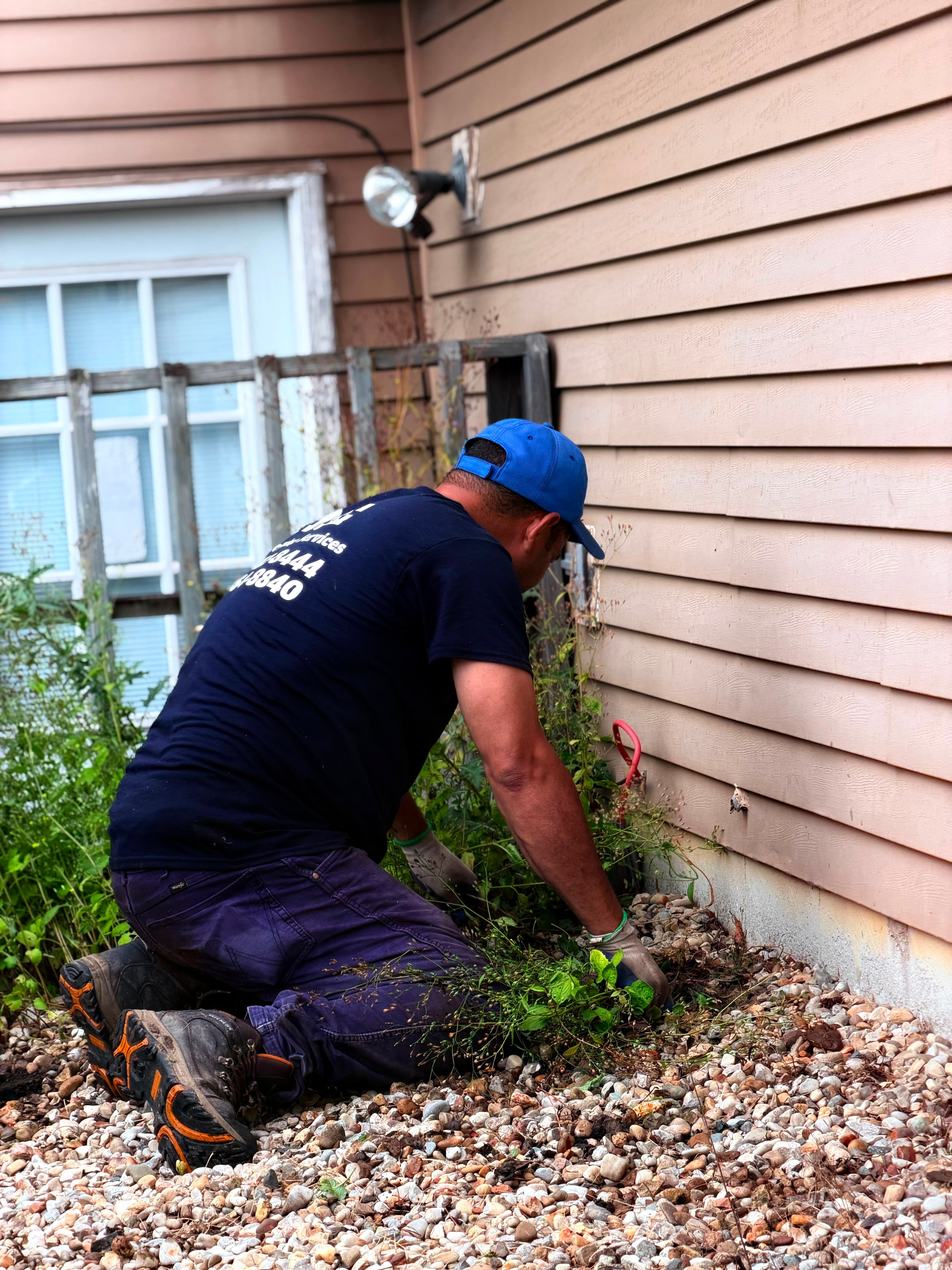 A man in a blue cap kneels on gravel, weeding beside a beige house.