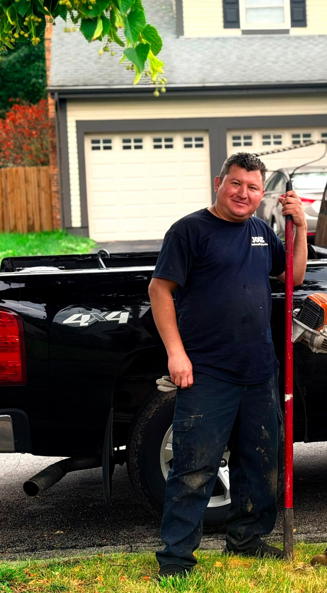 Smiling man in work clothes stands beside a black pickup truck holding a red tool.